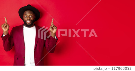 Winter holidays and shopping concept. Cheerful african american man in party outfit pointing fingers up, showing logo and smiling happy, red background 117963052