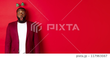 Merry Christmas. Happy african american man celebarting New Year, wearing party headband and making funny faces , standing over red background Merry Christmas. Happy african american man celebarting New Year, wearing party headband and making funny faces , standing over red background 117963087