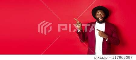 Winter holidays and shopping concept. Happy Black man pointing left and smiling, showing new year promo offer on red background Winter holidays and shopping concept. Happy Black man pointing left and smiling, showing new year promo offer on red background 117963097