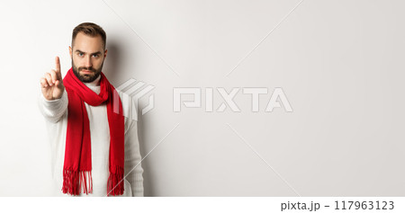 Serious man saying no, showing stop sign, shaking finger in decline, rejection sign, prohibit action, standing in winter sweater and red scarf against white background Serious man saying no, showing stop sign, shaking finger in decline, rejection sign, prohibit action, standing in winter sweater and red scarf against white background 117963123