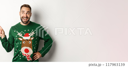New year celebration and winter holidays concept. Dreamy bearded man in green christmas sweater, pointing at upper left corner and smiling amused, white background 117963139