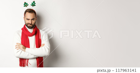 Christmas party and celebration concept. Suspicious young bearded man looking doubtful, wearing funny accessory hat, standing against white background 117963141