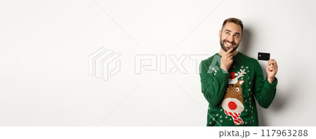 Christmas holidays and shopping concept. Thoughtful man planning New Year gift list, holding credit card and smiling, standing over white background 117963288