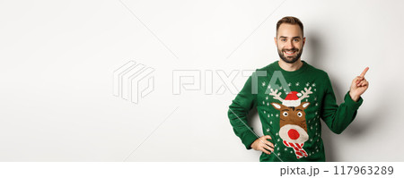 Winter holidays and christmas. Handsome bearded adult man pointing finger right at promo banner, standing in funny sweater against white background Winter holidays and christmas. Handsome bearded adult man pointing finger right at promo banner, standing in funny sweater against white background 117963289