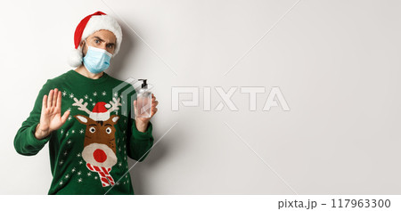 Concept of covid-19 and Christmas holidays. Man in face mask and santa hat stop person, asking to use hand sanitizer, standing over white background Concept of covid-19 and Christmas holidays. Man in face mask and santa hat stop person, asking to use hand sanitizer, standing over white background 117963300