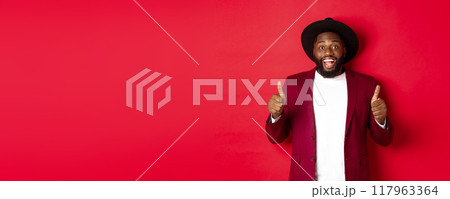 Christmas shopping and people concept. Handsome Black man smiling satisfied, showing thumbs-up, like and agree, approve something, standing against red background Christmas shopping and people concept. Handsome Black man smiling satisfied, showing thumbs-up, like and agree, approve something, standing against red background 117963364