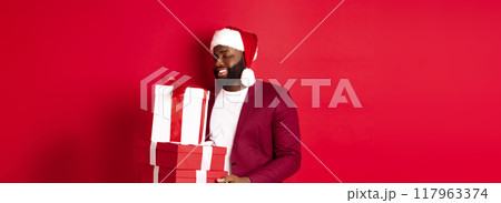 Christmas, New Year and shopping concept. Happy african amaerican man looking touched and thankful at xmas presents, receiving gifts, wearing santa hat, red background Christmas, New Year and shopping concept. Happy african amaerican man looking touched and thankful at xmas presents, receiving gifts, wearing santa hat, red background 117963374