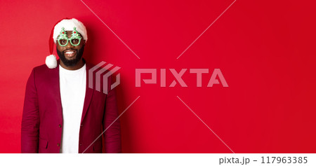 Funny Black man in santa hat and party glasses celebrating Christmas, smiling happy and wishing merry xmas, standing over red background Funny Black man in santa hat and party glasses celebrating Christmas, smiling happy and wishing merry xmas, standing over red background 117963385