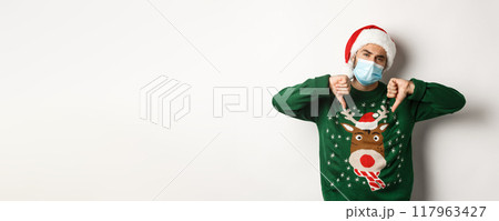 Concept of covid-19 and Christmas holidays. Displeased guy in face mask and santa hat showing thumbs down, standing over white background Concept of covid-19 and Christmas holidays. Displeased guy in face mask and santa hat showing thumbs down, standing over white background 117963427