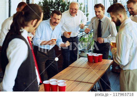 Businessmen, colleagues taking break, playing beer pong at the bar, mixing work with playful and joyful time. 117963718