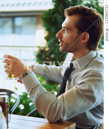 Businessman in formal wear sitting at table at local bar with glass of lager beer and talking communicating with business partners in informal settings Businessman in formal wear sitting at table at local bar with glass of lager beer and talking communicating with business partners in informal settings 117963722