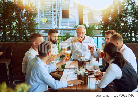 Productive meeting, businessmen in formal wear gathering at outdoor bar, drinking beer, talking, laughing and relaxing after hard working week 117963740