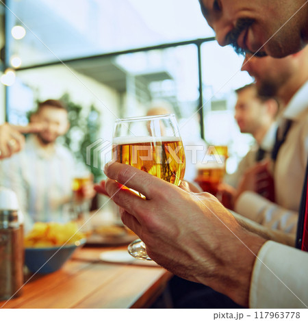 Focus on male hand holding lager beer. Business people, colleagues meeting at local bar, drinking beer, talking and celebrating successful collaboration Focus on male hand holding lager beer. Business people, colleagues meeting at local bar, drinking beer, talking and celebrating successful collaboration 117963778