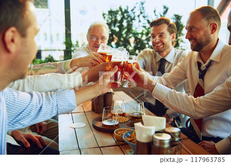 Group of cheerful men, business people meeting at outdoor cafe, clinking glasses with lager beer and celebrating successful deal. 117963785