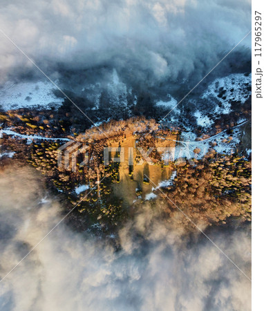 Aerial view capturing winding path through snowy, mist-covered landscape. Contrast between snow-dusted terrain and enveloping clouds creates dramatic and ethereal scene, highlighting beauty of nature. 117965297