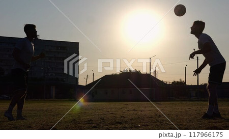 Silhouette of professional footballers kicking soccer ball with their heads on stadium at sunset. Two sportsmen showing tricks with ball while passing it to each other at field. Freestyle football 117966175