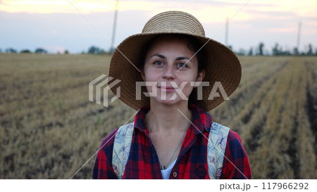 Happy smiling female farmer looks into camera standing at wheat field. Portrait of young beautiful agronomist in straw hat with barley meadow at background. Agricultural business concept 117966292