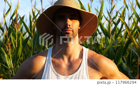 Serious male farmer looks into camera standing near corn field. Portrait of young handsome agronomist in straw hat with maize meadow at background. Concept of agricultural business. Close up 117966294
