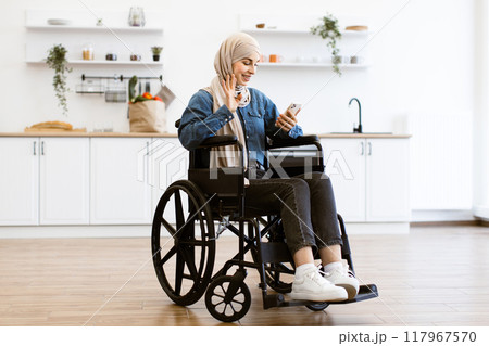 Muslim woman in wheelchair using smartphone at home kitchen Muslim woman in wheelchair using smartphone at home kitchen 117967570