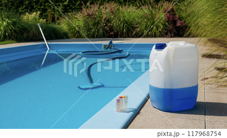 Pool cleaning hose and vacuum head with a large chemical container and water testing kit by the poolside, emphasizing maintenance and water quality. Pool cleaning hose and vacuum head with a large chemical container and water testing kit by the poolside, emphasizing maintenance and water quality. 117968754