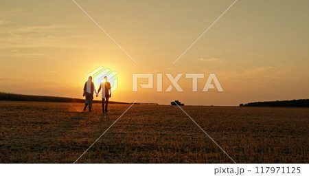 A couple of farmers walk hand in hand through a field at sunset, with a combine harvester in the distance. A couple of farmers walk hand in hand through a field at sunset, with a combine harvester in the distance. 117971125