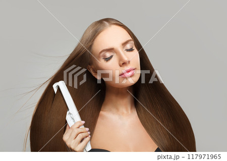 Pretty brunette woman holding straight hair iron and straightening her long smooth shiny hair 117971965