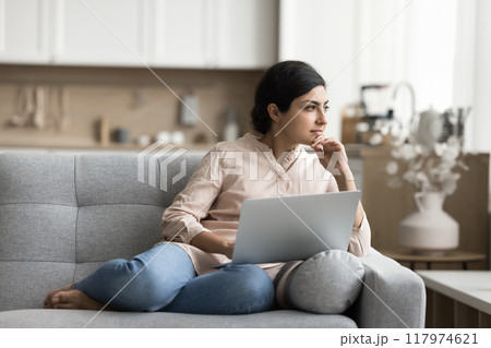Pensive woman studying or working seated on sofa with laptop Pensive woman studying or working seated on sofa with laptop 117974621