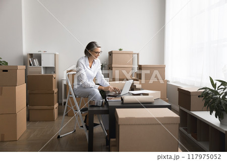 Woman working on laptop in room filled with cardboard boxes 117975052
