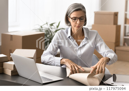 Serious mature woman wrapping parcel with paper 117975094