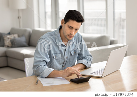 Serious young Latin homeowner man calculating domestic expenses Serious young Latin homeowner man calculating domestic expenses 117975374
