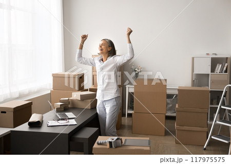 Happy mature woman standing in stockroom filled with cardboard boxes Happy mature woman standing in stockroom filled with cardboard boxes 117975575