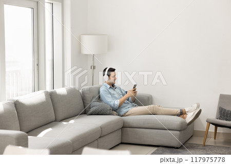 Lazy young Latin man in headphones resting on comfortable couch Lazy young Latin man in headphones resting on comfortable couch 117975778