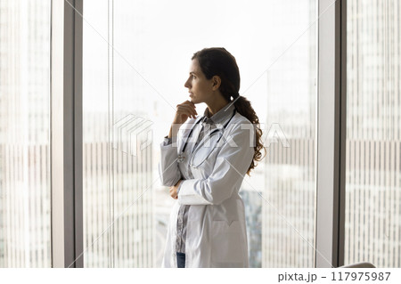 Thoughtful female doctor gazing out of window with cityscape view 117975987