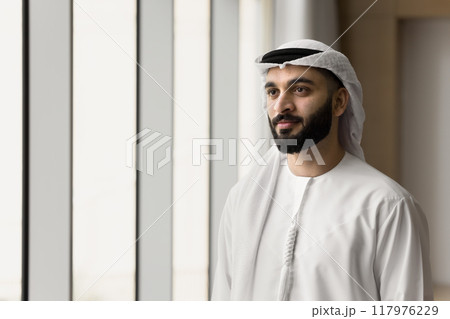 Positive handsome young Arabic businessman standing at office window Positive handsome young Arabic businessman standing at office window 117976229