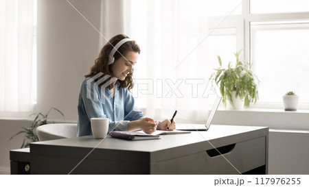 Focused young woman in headphones studying at home using laptop Focused young woman in headphones studying at home using laptop 117976255