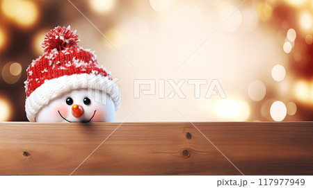 Two adorable snowman peek over a wooden fence, wearing cozy knit hats against a backdrop of golden holiday lights. The warm bokeh effect creates a festive, cheerful atmosphere, ideal for Christmas 117977949