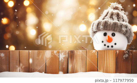 Two adorable snowman peek over a wooden fence, wearing cozy knit hats against a backdrop of golden holiday lights. The warm bokeh effect creates a festive, cheerful atmosphere, ideal for Christmas 117977950