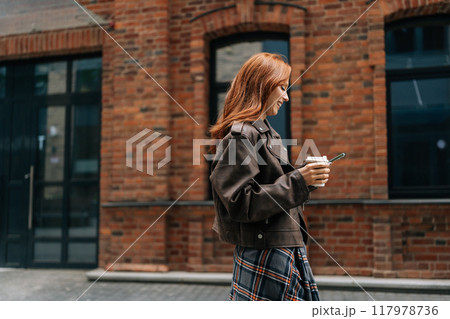 Side view of cheerful young woman in brown jacket walking down street and using smartphone, smiling looking to screen. Stylish redhead female going typing mobile phone enjoying free time outdoors. 117978736