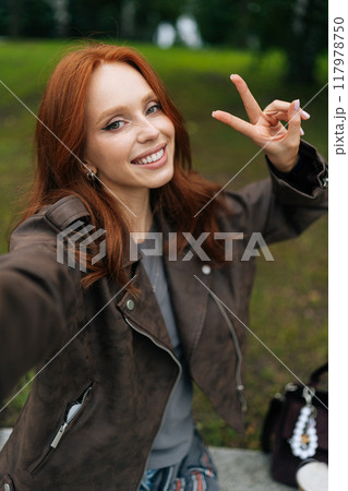 Vertical portrait of positive young woman with long red hair smiling happily looking at camera, taking selfie in park, showing peace sign. Happy redhead female shooting selfie flashing peace sign. 117978750