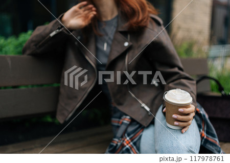 Cropped shot of unrecognizable woman wearing brown leather jacket and jeans sitting on bench outside, holding cardboard cup of coffee, savoring moment of relaxation and fashion in city. Cropped shot of unrecognizable woman wearing brown leather jacket and jeans sitting on bench outside, holding cardboard cup of coffee, savoring moment of relaxation and fashion in city. 117978761