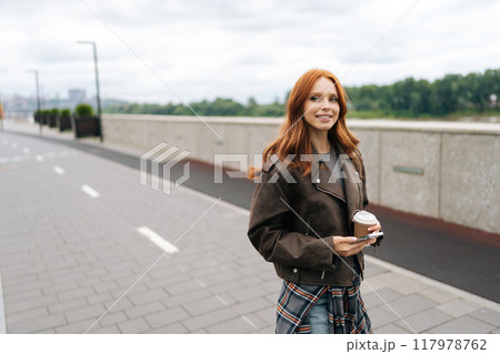 Smiling young woman enjoying stroll along riverfront promenade, holding takeaway coffee cup on cloudy day. Happy redhead female in leather jacket enjoying peaceful moment with coffee. Smiling young woman enjoying stroll along riverfront promenade, holding takeaway coffee cup on cloudy day. Happy redhead female in leather jacket enjoying peaceful moment with coffee. 117978762