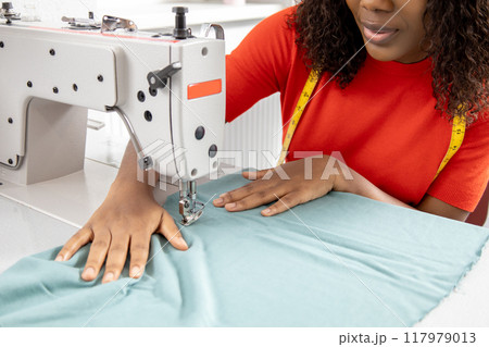 African American woman sewing clothes from light blue textile with sewing machine African American woman sewing clothes from light blue textile with sewing machine 117979013