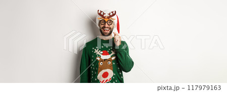 Christmas and holidays concept. Happy bearded man celebrating xmas, holding party mask and wearing Santa hat, smiling amazed, standing over white background Christmas and holidays concept. Happy bearded man celebrating xmas, holding party mask and wearing Santa hat, smiling amazed, standing over white background 117979163