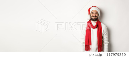 Christmas holidays. Bearded man in santa hat and red scarf looking happy, enjoying New Year party celebration, standing over white background 117979250