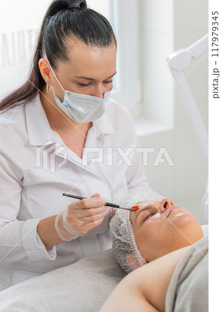 Beautician applying peeling moisturizing cream mask on young woman face for rehydrate face skin 117979345