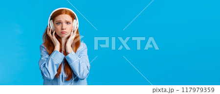 Girl about cry from emotional song. Upset redhead girlfriend suffering breakup, sobbing listening music in large white headphones, standing blue background in nightwear, unhappy Girl about cry from emotional song. Upset redhead girlfriend suffering breakup, sobbing listening music in large white headphones, standing blue background in nightwear, unhappy 117979359