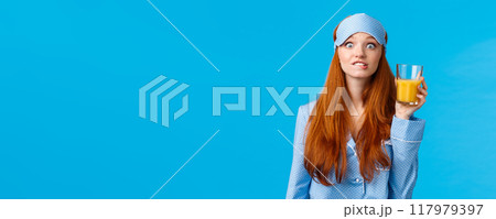 Girl likes drinking fresh juices. Cheerful and cute pretty feminine ginger girl, foxy teenager in nightwear, sleep mask, biting lip eager, seeing something tempting, holding glass orange juice 117979397