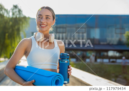 Happy young sportswoman jogger with water bottle and rolled mat walking in city park on sunny day Happy young sportswoman jogger with water bottle and rolled mat walking in city park on sunny day 117979434
