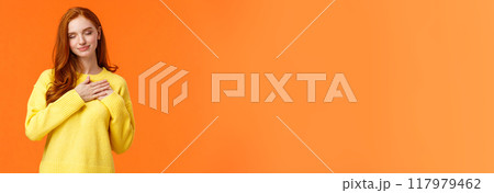 Tender and romantic, lovely young modern redhead woman 20s, touch heart and close eyes, remember dearest, touching moment, recall perfect date, keep memories in soul, orange background 117979462