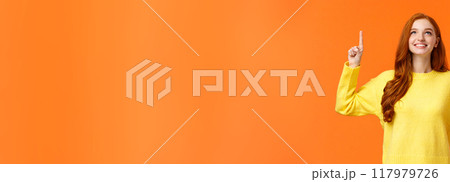 Dreamy happy and upbeat redhead young modern woman gazing up with satisfied smile. Gorgeous girl ginger hair conemplate top advertisement, pointing upwards, standing orange background 117979726